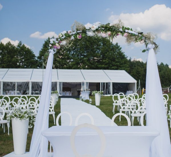 Freiluft-Alea für Trauung mit Blumenbogen, weißen Stühlen und Festzelt auf Rasenfläche unter blauem Himmel
