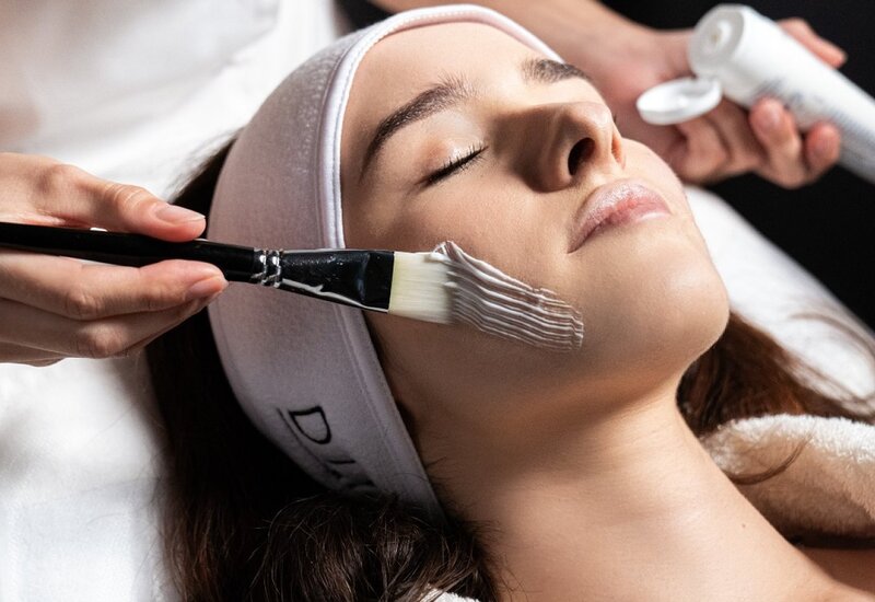 Guest reclining in spa treatment room, eyes closed as esthetician brushes a white facial mask from a tube; calm, restorative ambiance.