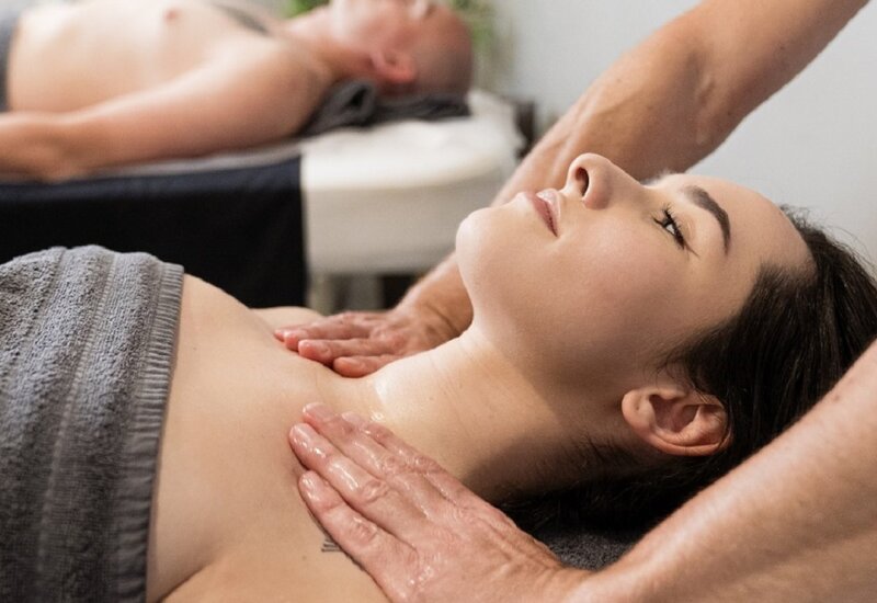 Couples spa massage: woman reclines on a treatment bed as therapist oils and massages her neck and chest, gray towel