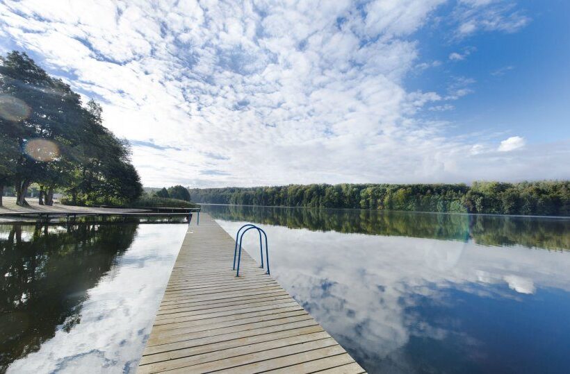 Drewniany pomost nad jeziorem z drabinką do wody, spokojna lustrzana tafla, zalesiony brzeg i błękitne niebo.