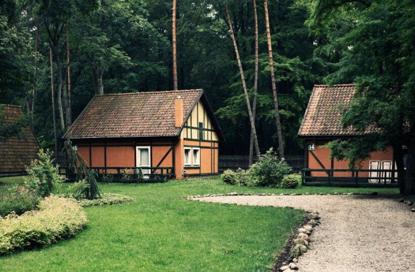 Szachulcowe domki letniskowe z dachówką w sosnowym lesie, z trawnikiem i żwirową ścieżką.