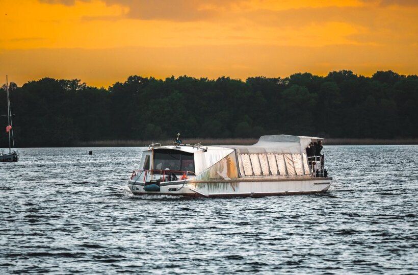 Łódź wycieczkowa o zachodzie słońca z zabudowaną kabiną i odkrytym tylnym pokładem, pasażerowie, płynie po spokojnym jeziorze
