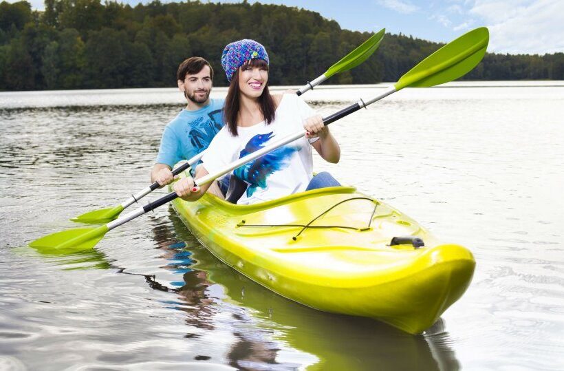 Para w jasnożółtym kajaku dwuosobowym na spokojnym jeziorze z zalesionym brzegiem — hotelowa wypożyczalnia kajaków.