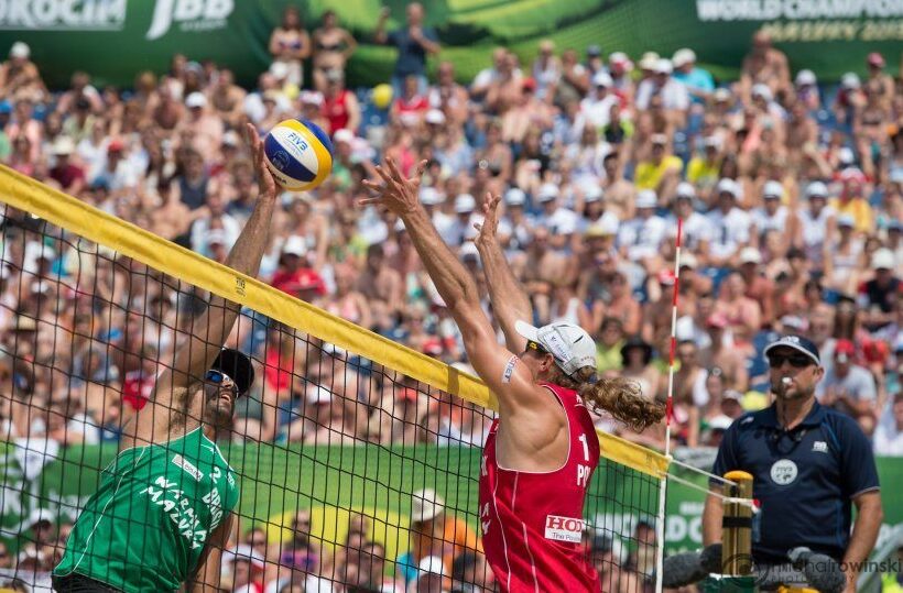 Spannendes Beachvolleyball am Netz, Spieler in Rot blockt den Ball, Schiedsrichter und dicht gefüllte Zuschauertribünen