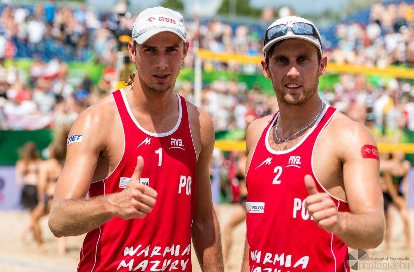 Zwei Beachvolleyballspieler in roten Polen‑Trikots zeigen Daumen hoch auf sandigem Spielfeld vor Zuschauern