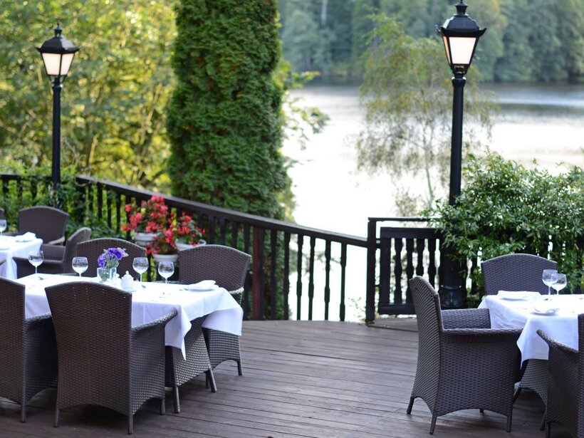 Seeseitige Restaurantterrasse mit Rattanstühlen, weiß gedeckten Tischen, Glasgeschirr, blühenden Kübelpflanzen, Holzdeck und Laternen