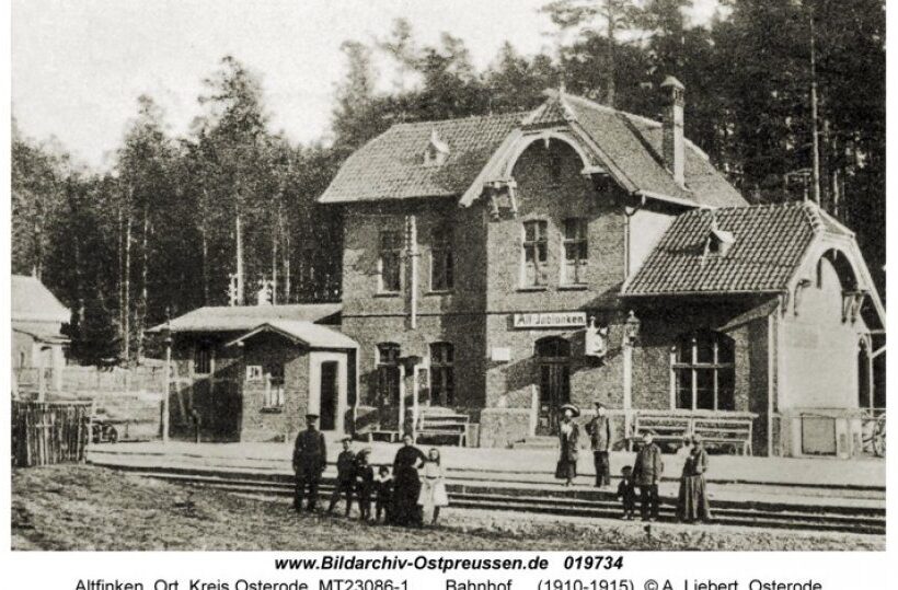 Historischer Ziegelbahnhof mit Ziegeldach und geschwungenen Giebeln, Bahnsteig mit Reisenden, Wald im Hintergrund.
