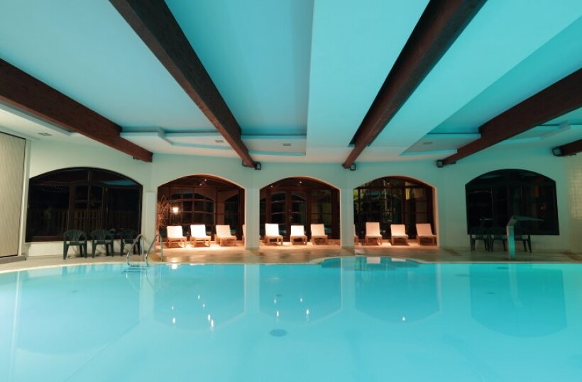 Hallenbad im Hotel mit türkisblauem Wasser, sichtbaren Holzträgern, rundbögigen Fenstern und Liegestühlen.