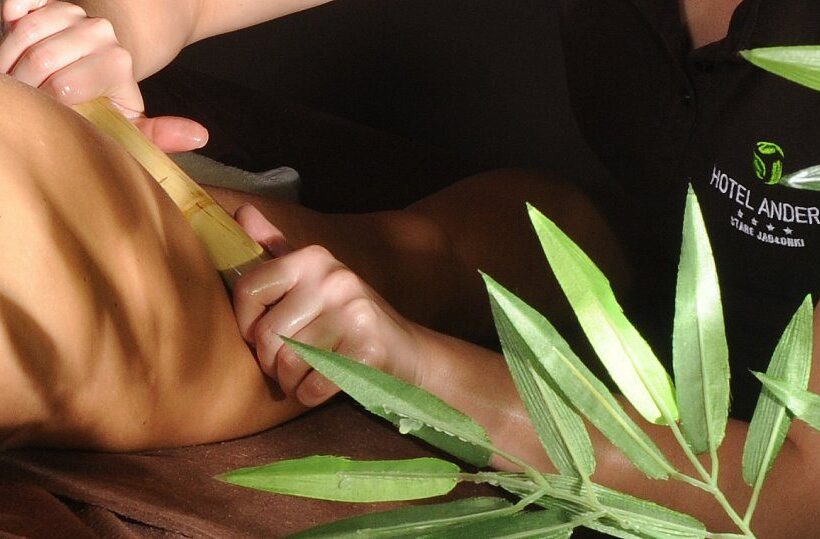 Therapeut führt Bambusmassage am Rücken eines Gastes durch, grüne Blätter im Bild, entspannende Hotel-Spa-Behandlung.