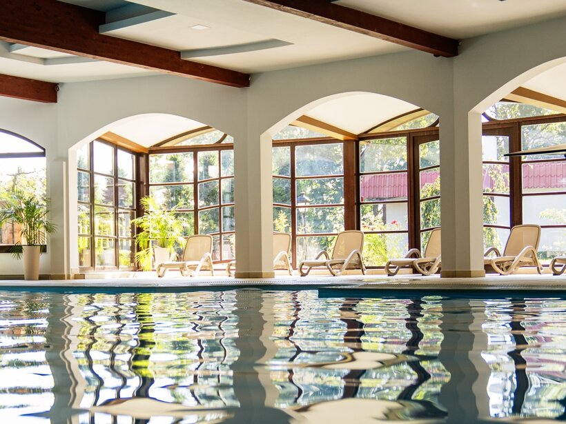 Indoor hotel pool with arched floor-to-ceiling windows, sunlit loungers, potted plants and exposed wooden ceiling beams