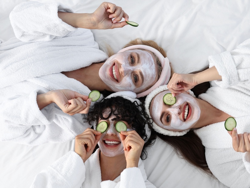 Drei Gäste in weißen Bademänteln liegen auf dem Bett mit Gesichtsmaske und Gurkenscheiben und genießen eine Spa-Behandlung im Zimmer.