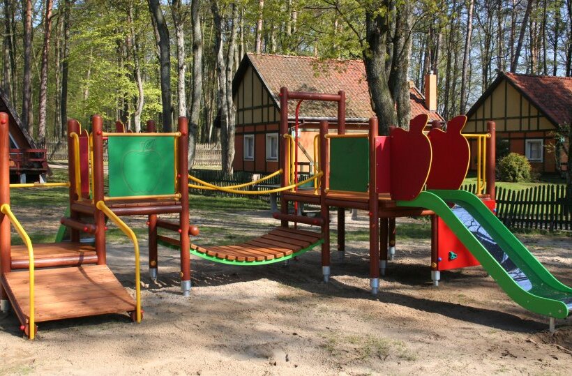 Children's playground with green slide, wooden climbing bridge and play panels beside timber cottages in a shady forested resort yard