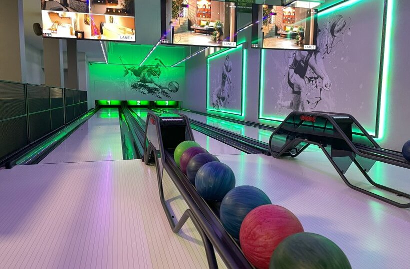 Modern hotel bowling alley with neon green lighting, two lanes, colorful bowling balls and large sports murals for lively guest entertainment.