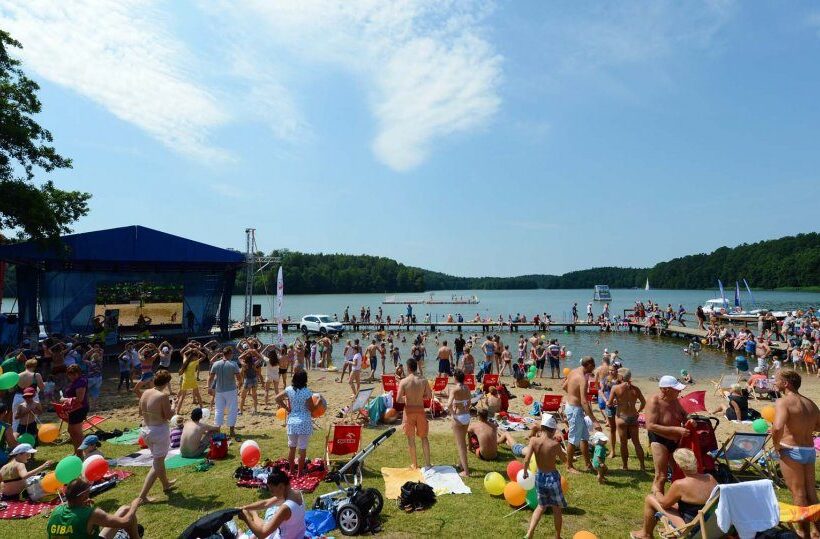Busy lakeside family festival with stage and live music, sandy beach and swimming pier, sunlit forested shoreline and colorful balloons.