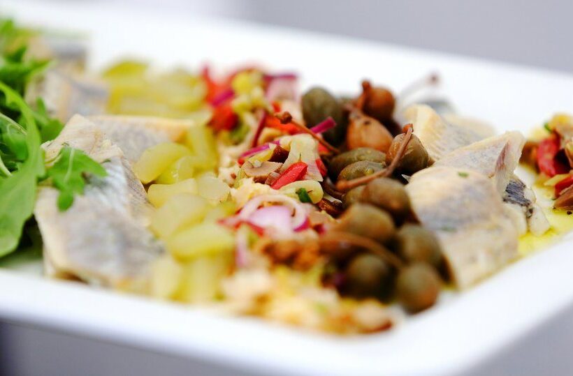 Marinated herring fillets with capers, diced pickles, red onion and arugula on a white plate — cold seafood appetizer.