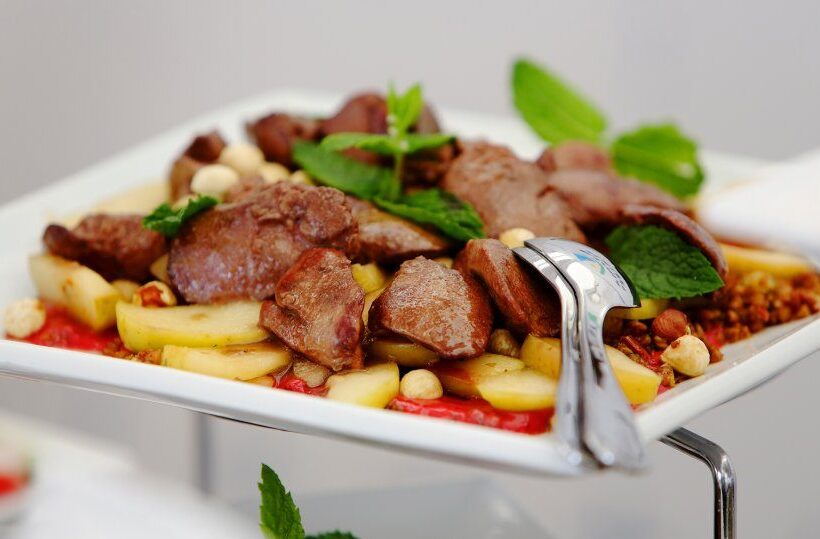 Roasted meat medallions on sautéed potato slices with hazelnuts, mint garnish and serving tongs on a white buffet platter.