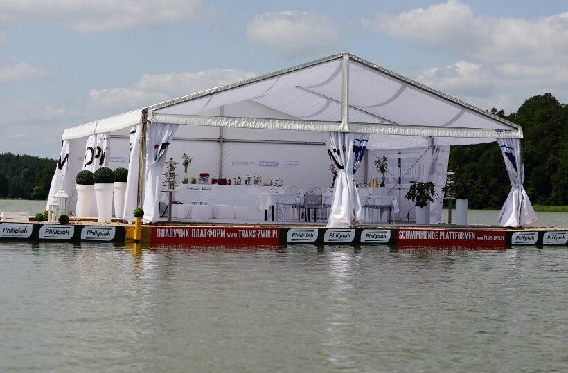 White open-sided event tent on a floating platform over a calm lake, set with a buffet, dining tables, white chairs and potted plants