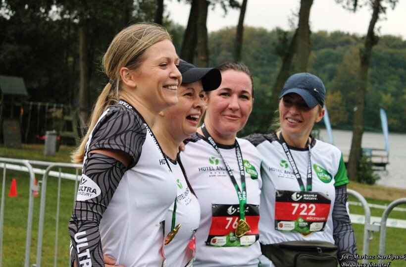 Four smiling female runners in sponsor shirts with medals, arm‑in‑arm at a lakeside race finish surrounded by trees.
