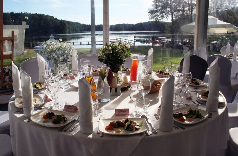 Lakeside wedding reception table with white linens, floral centerpiece, folded napkins, plated starters and glassware overlooking a wooden jetty.