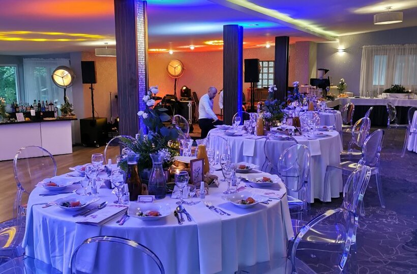 Banquet room with round tables in white linen, floral centerpieces, clear chairs, colorful uplighting and staff preparing service.