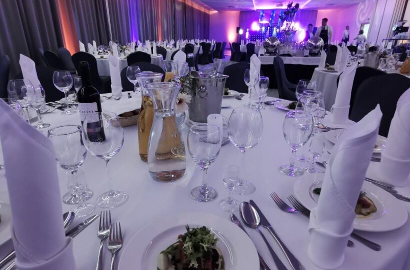 Banquet hall set for gala: round tables with white linens, folded napkins, wine, glassware, starters and purple lighting.