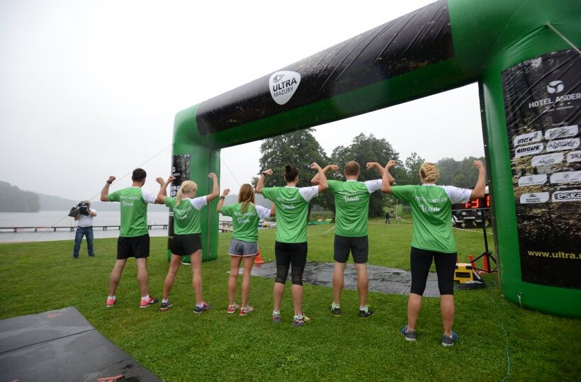 Team in matching green shirts flexing under a lakeside finish arch during an outdoor team-building run