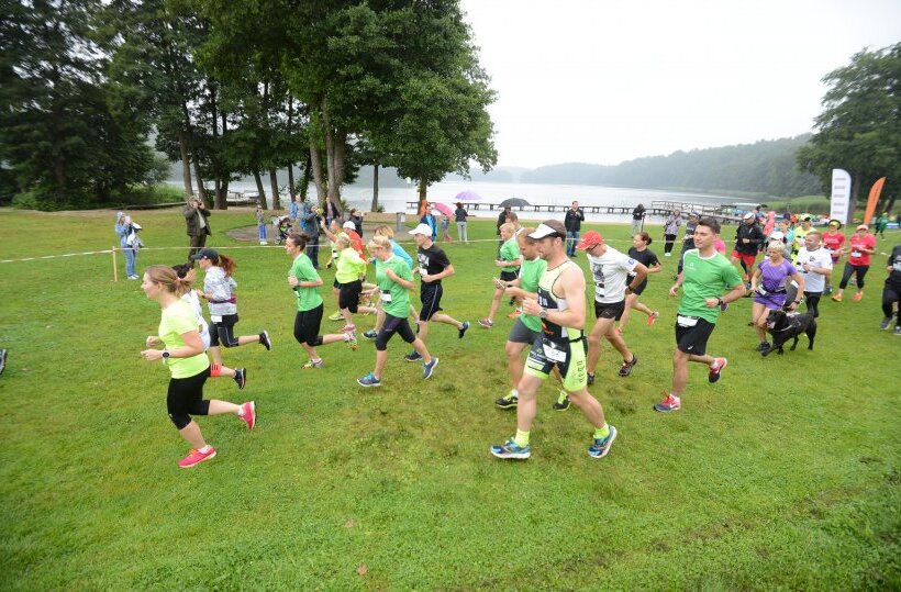 Runners in bright shirts start a lakeside fun run across a grassy park with trees and wooden docks by a misty lake
