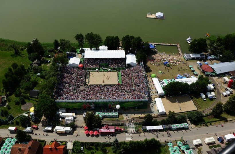Lakeside beach volleyball stadium with packed grandstand, sandy courts, vendor tents and a lively beachside festival atmosphere.