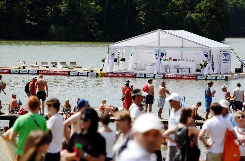 Floating white event tent and lounge deck on a lake, sunbeds and guests relaxing on the shore during a summer festival