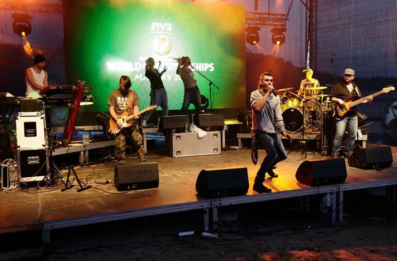Live band performing on an illuminated outdoor stage with LED backdrop, singer fronting, guitar, bass, drums and energetic stage lighting.