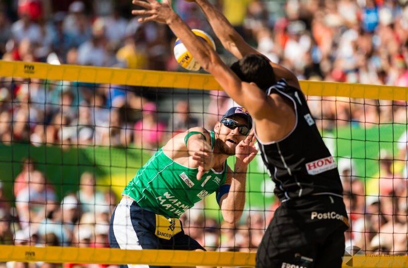 Two beach volleyball players at the net; player in green spikes while opponent blocks, packed sunny stadium crowd.