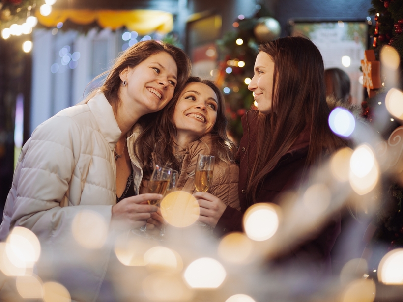 Three women in winter coats clinking champagne amid twinkling holiday lights and festive decorations, smiling together.