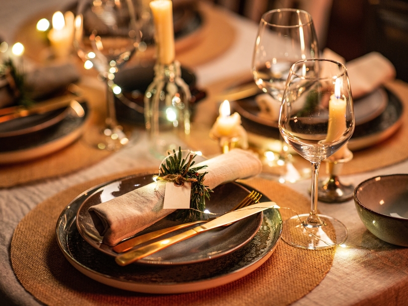 Warm, kerzenbeleuchtetes Tischgedeck mit Keramiktellern, goldenem Besteck, Leinenserviette mit Zweig und Weinglas