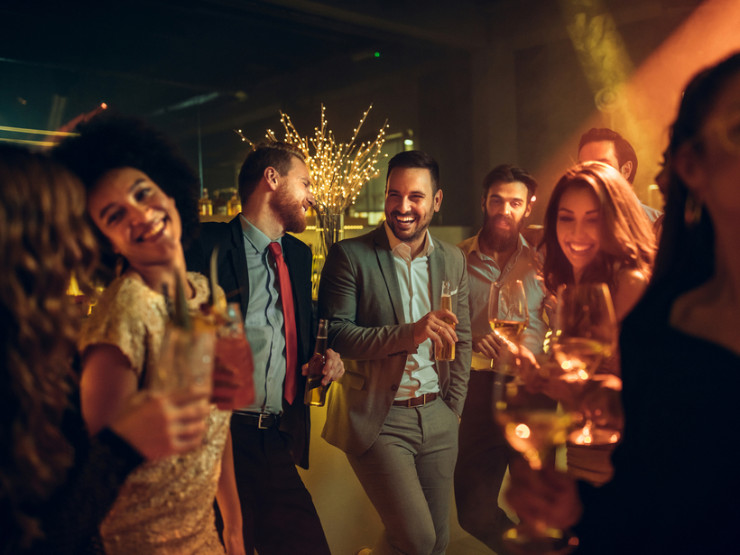 Lebhafte Hotel-Cocktailparty in gedämpfter Lounge — Gäste in Abendgarderobe lachen und halten Wein, Cocktails und Bier an der stimmungsvoll beleuchteten Bar.