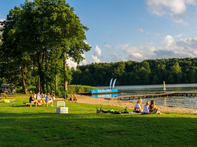 Seeufer mit Picknickwiese, Holzbänken, Sandstrand, Steg und Gästen, die im Schatten der Bäume entspannen.