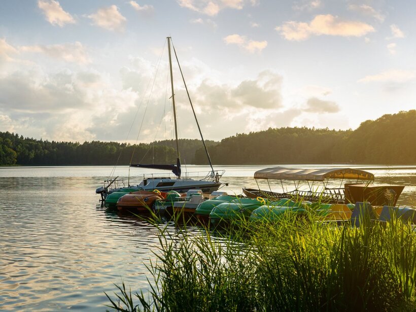 Seeseitiger Steg bei Sonnenuntergang mit vertäutem Segelboot, überdachten Ruderbooten und bunten Tretbooten, ruhiges Wasser und bewaldetes Ufer