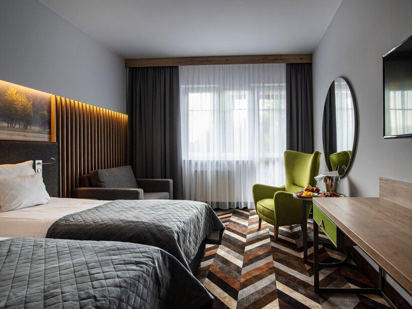 Nowoczesny pokój hotelowy z dwoma łóżkami, podświetlanym drewnianym wezgłowiem, zielonym fotelem, biurkiem, telewizorem oraz owocami i wiaderkiem na szampana