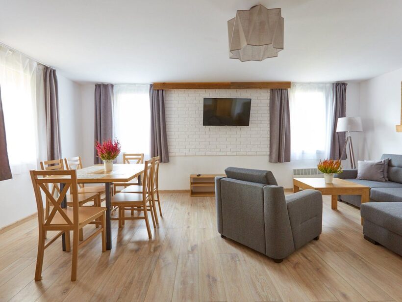 Jasny, nowoczesny salon w apartamencie z drewnianym stołem (6 krzeseł), szarą sofą i fotelem, telewizorem na ścianie i doniczkami z kwiatami