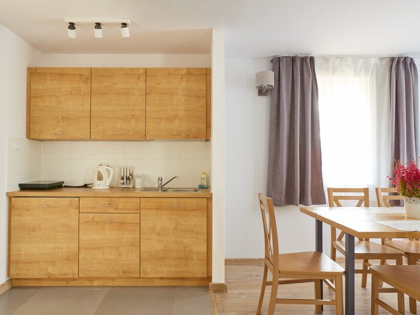 Kuchnia hotelowa z drewnianymi szafkami, zlewem, czajnikiem i płytą; stół z krzesłami i bukietem przy jasnym oknie.