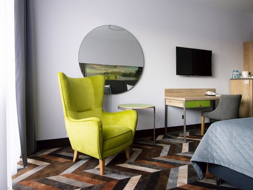 Jasny nowoczesny pokój z limonkowym fotelem, okrągłym lustrem, biurkiem z TV, czajnikiem i pikowanym łóżkiem