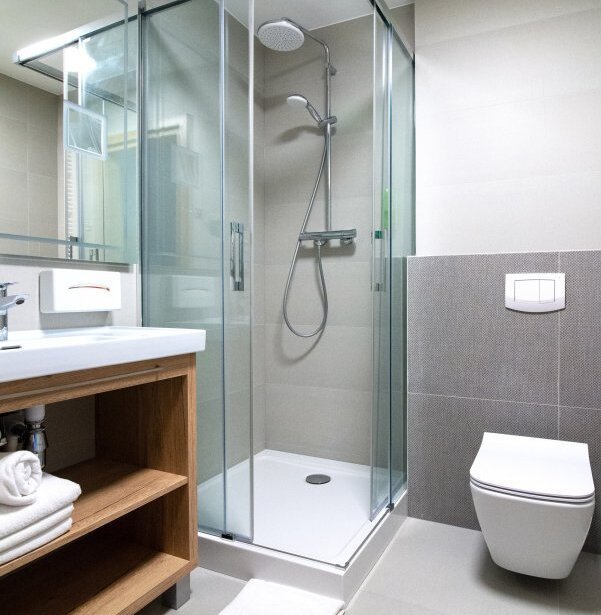 Nowoczesna łazienka hotelowa z przeszklonym prysznicem i deszczownicą, drewniana szafka z ręcznikami i wisząca toaleta