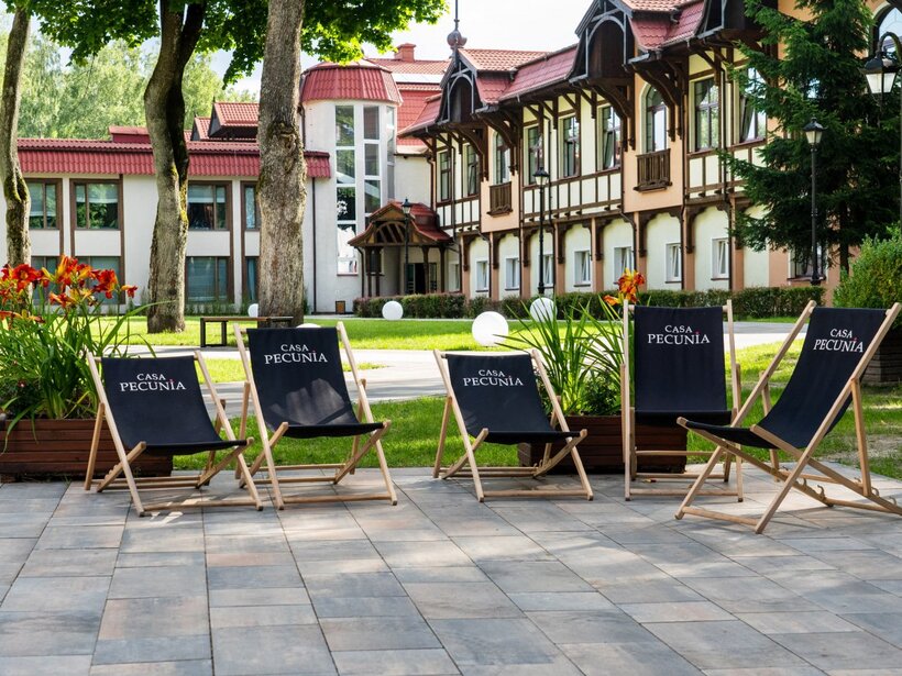 Casa Pecunia Liegestühle auf gepflasterter Terrasse, neben Rasen, Bäumen und blühenden Kübeln vor einem Hotel mit Holzfassade.