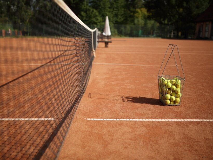 Roter Sandplatz mit Netz im Vordergrund, Drahtkorb voller gelber Bälle an der Grundlinie, Bank und Sonnenschirm im Hintergrund
