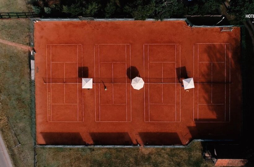 Aerial view of four clay tennis courts at the hotel, three white sun umbrellas, players and shaded sidelines on grassy grounds
