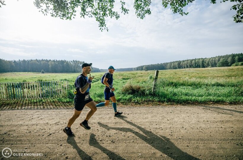 Two runners on a sunlit rural dirt trail beside green fields and a distant tree line, active outdoor run