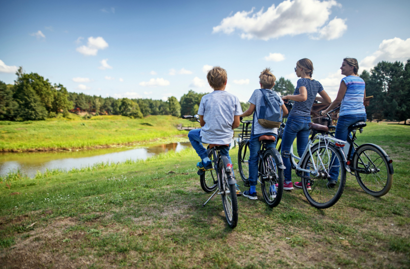 Family of four on bicycles paused by a calm riverside meadow under a sunny blue sky.