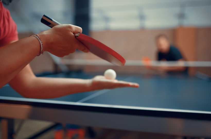 Guest serving a ping-pong ball at an indoor table in a hotel's game room, paddle poised, opponent blurred across the table.