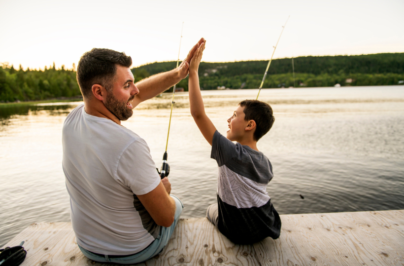 Father and son high-five on a wooden lakeside dock while fishing at sunset on the hotel waterfront.