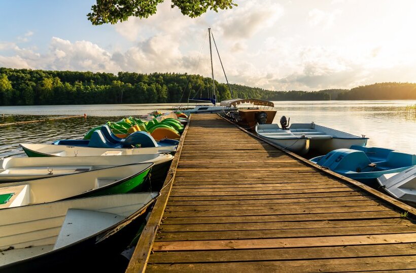 Wooden lakeside dock with rental rowboats, colorful pedal boats and a sailboat at sunset over calm water.