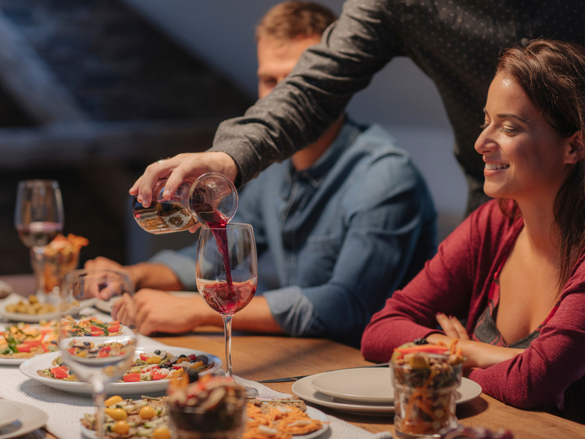 Person pouring red wine into glass at a cozy table with shared appetizers; smiling woman enjoys relaxed dinner ambiance.